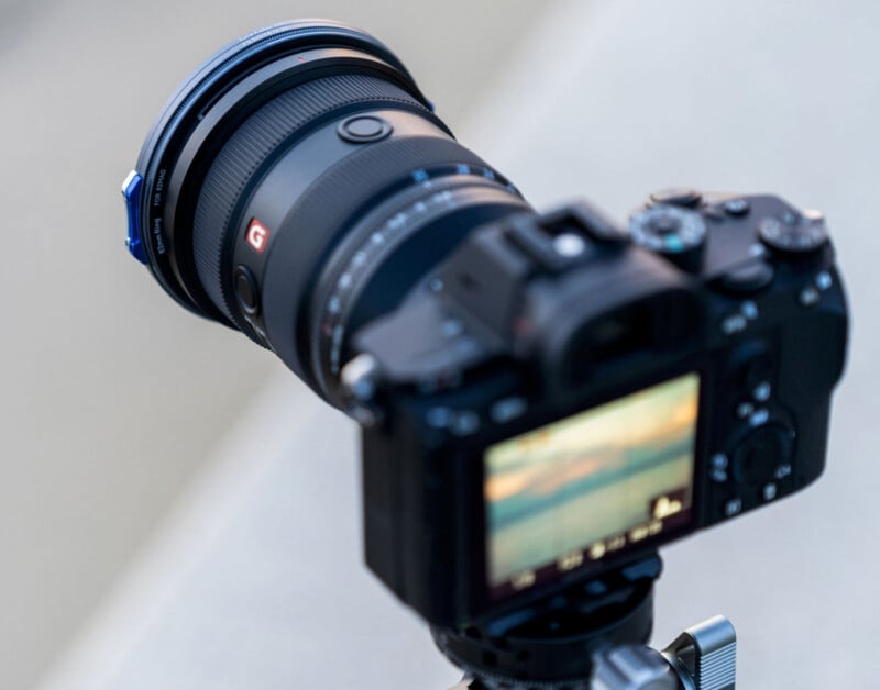 A close-up of a DSLR camera mounted on a tripod, focused on its lens. The camera's display screen shows a blurred image of a sunset or landscape. The lens has a distinct red "G" logo. The background is out of focus.