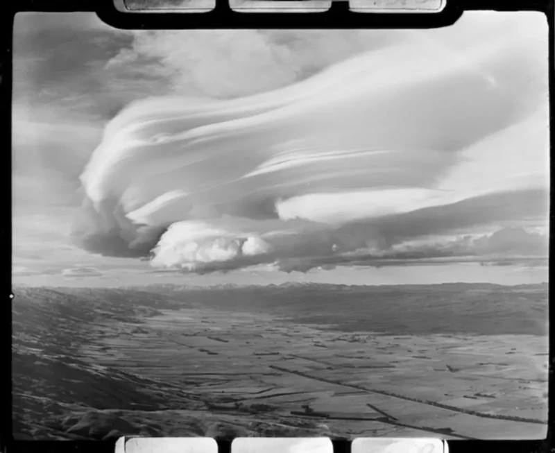 Black and white photo of a vast landscape with dramatic, swirling clouds in the sky. The ground is a patchwork of fields bordered by mountains in the distance, creating a striking contrast with the dynamic cloud formation above.