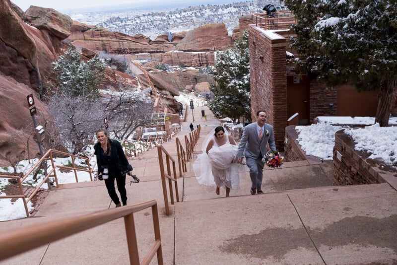 A bride and groom in wedding attire walk up snowy steps at an outdoor venue. The bride holds her dress, and the groom carries a bouquet. A photographer follows them, capturing the moment. Snow-covered rocks and trees surround the scene.