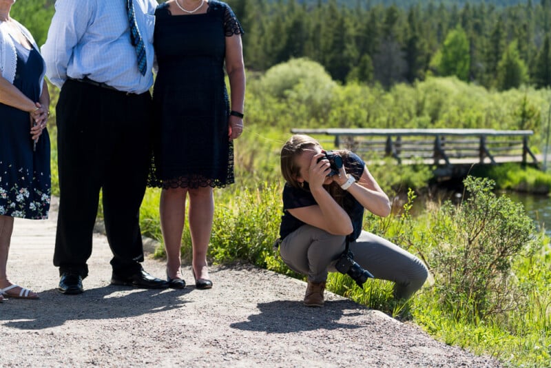 A photographer crouches on a gravel path taking a picture of three people standing nearby. They are outdoors near a green park with trees and a small bridge in the background on a sunny day.