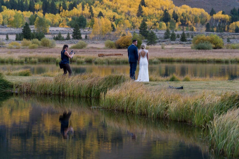 A photographer captures a couple in wedding attire standing by a reflective lake. The background features vibrant autumn trees with yellow leaves. The scene is peaceful, with a natural landscape of grass and distant mountains.