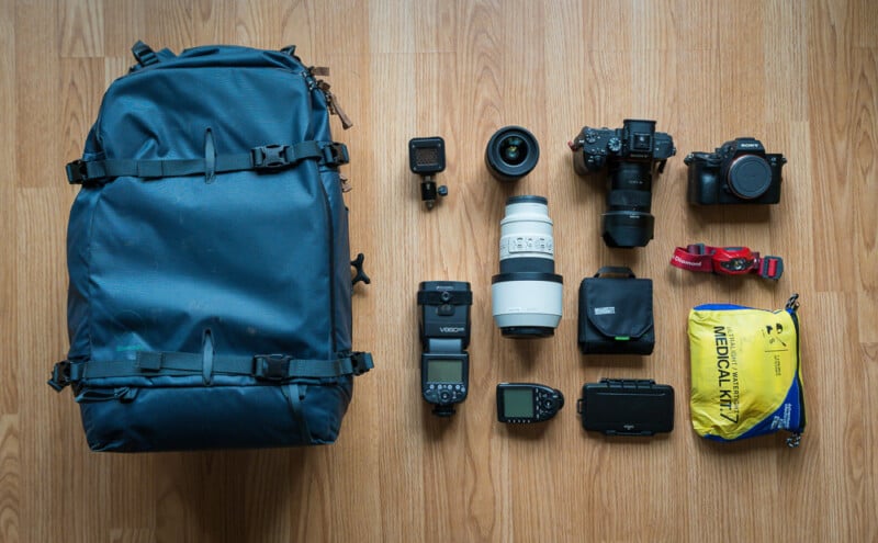 A blue backpack sits on a wooden floor surrounded by photography gear: two cameras with lenses, a zoom lens, a flash, a video light, a GoPro, a GPS device, a memory card wallet, a headlamp, and a yellow medical kit.