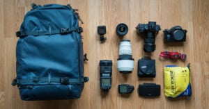 A blue backpack is laid on a wooden floor next to various camera equipment, including two cameras with lenses, a zoom lens, a flash, a small action camera, batteries, and a yellow medical kit.