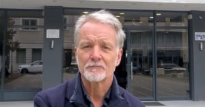 A man with grey hair and a beard stands outside in front of a glass building entrance, wearing a dark jacket. The street in the background has a few parked cars.