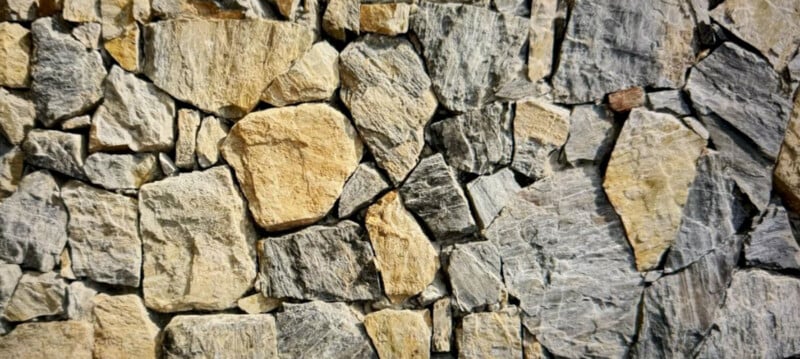 A close-up of a rustic stone wall made up of different sized and shaped stones. The stones vary in color from light beige to dark gray and are joined together with uneven joints, creating a rough, natural texture.