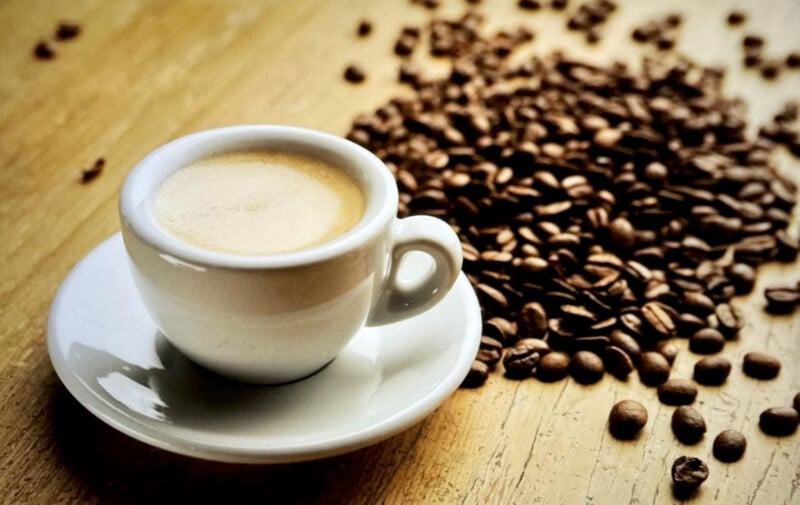 A white ceramic cup filled with espresso sits on a saucer on a wooden table. A pile of roasted coffee beans is scattered around the cup, creating a warm and inviting atmosphere.