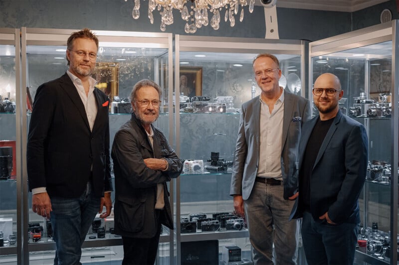 Four men in suits and jackets stand in front of glass display cases filled with cameras. A chandelier hangs above them, and the setting has a vintage and elegant atmosphere.