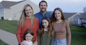 A family of five stands outside their home, smiling at the camera. The group includes a man, a woman, two older girls, and a younger girl. They are dressed in casual clothing and are positioned in front of suburban houses with a lawn in the background.