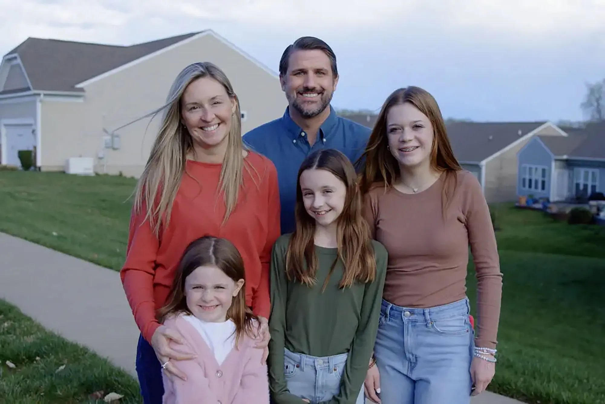 A family of five stands outside in front of houses. The parents are in the back, with the mother wearing a red top and the father in a blue shirt. The three daughters in front wear green, pink, and brown tops. They all smile at the camera.