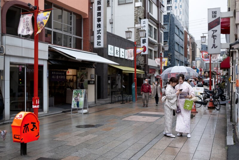 People in traditional attire stand under an umbrella on a rainy street in a Japanese city. They are looking at something, possibly a map or phone. The street is lined with shops and signs, and a bright orange mailbox is in the foreground.