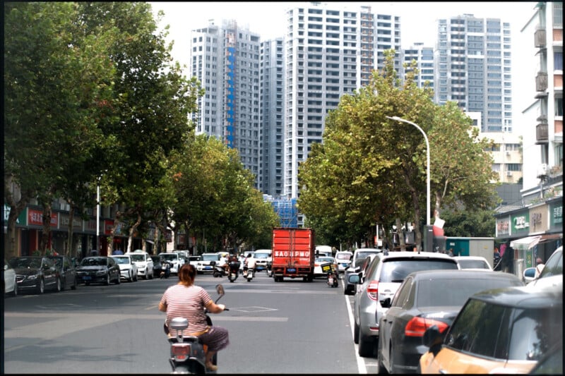 A city street with parked cars along both sides. A person rides a scooter in the foreground, approaching a red truck. Tall residential buildings and lush green trees line the street. The sky is partially cloudy.