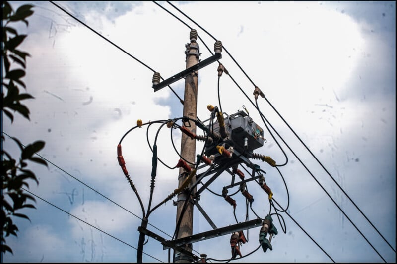 A wooden utility pole with a transformer and multiple power lines set against a partly cloudy sky. The pole is surrounded by wires and insulators, with some greenery visible in the lower left corner.