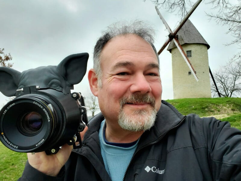 A smiling man holds a camera shaped like a pig's head near his face. In the background, a stone windmill with brown sails is set against a cloudy sky. He's wearing a black jacket and appears to be outdoors.