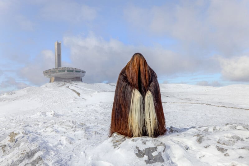 A figure wearing a long, flowing cloak with brown and white fur-like textures stands in a snowy landscape. In the background, a futuristic, abandoned building with a circular design and tall tower is visible under a partly cloudy sky.