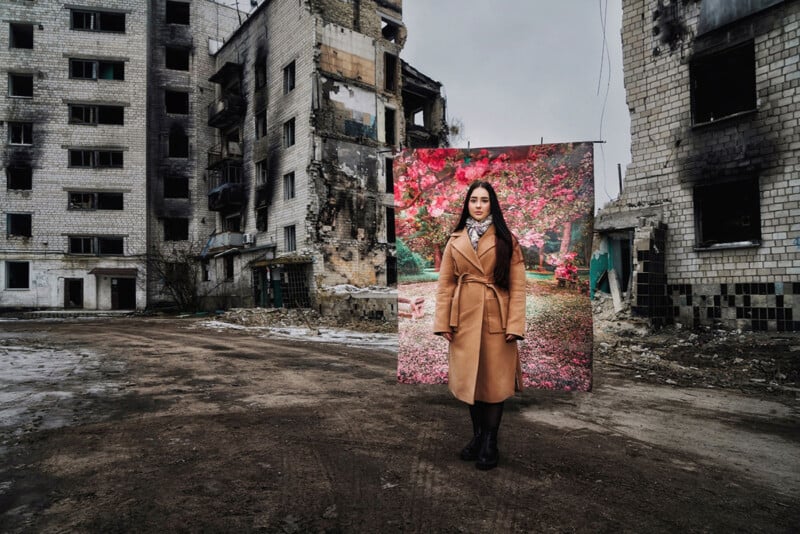 A woman stands in front of a backdrop showing a colorful flower scene amidst a desolate urban area with partially destroyed and weathered buildings. She is wearing a tan coat and black boots, and the contradiction between the backdrop and the surroundings is stark.