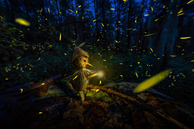 A small, whimsical doll made of natural materials stands on a log in a dark forest. It holds a tiny light, illuminating its face, while numerous fireflies glow around it, creating a magical, enchanting atmosphere.