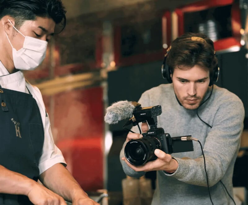 A person wearing a sweater and headphones holds a camera with a microphone, filming another person in an apron and mask who is preparing food. They appear to be in a kitchen or cooking studio.