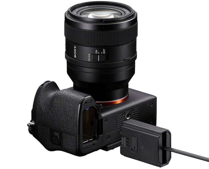 A digital camera with a detachable lens attached to a power adapter. The camera is turned upside down, showcasing its battery compartment. The cable connects from the adapter to the camera for charging or power supply.