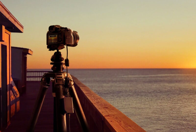 A camera on a tripod is set up on a wooden deck, facing a calm sea and orange-hued sunset. The sky is clear, and the horizon is visible in the background, capturing a serene moment by the water.