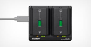 A Sony BC-QZ1 battery charger with two NP-FZ100 batteries inserted. Both batteries show green indicator lights for full charge. A charging cable is connected on the left side. The charger has a black design with labeled charging indicators.