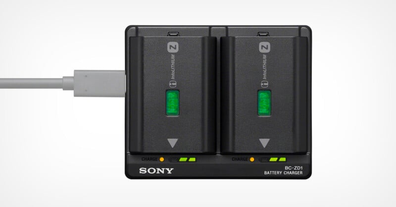 A Sony BC-QZ1 battery charger with two NP-FZ100 batteries inserted. Both batteries show green indicator lights for full charge. A charging cable is connected on the left side. The charger has a black design with labeled charging indicators.