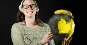 A person wearing glasses and a beanie smiles in front of a black background. They are holding a partially exploded banana, with banana pulp and peel visibly bursting outward. They're wearing a knitted sweater.