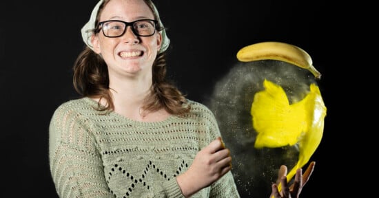 A person wearing glasses and a beanie smiles in front of a black background. They are holding a partially exploded banana, with banana pulp and peel visibly bursting outward. They're wearing a knitted sweater.