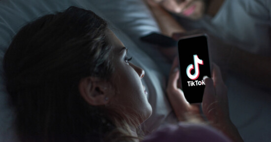 A woman lies in bed at night looking at her phone, which displays the TikTok logo. A blurred figure is using a phone in the background.