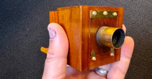 A hand holding a small wooden pinhole camera with brass fittings and a cylindrical lens. The camera has a simple, boxy design with a polished finish. The background is black, providing contrast to the camera's warm wood tones.