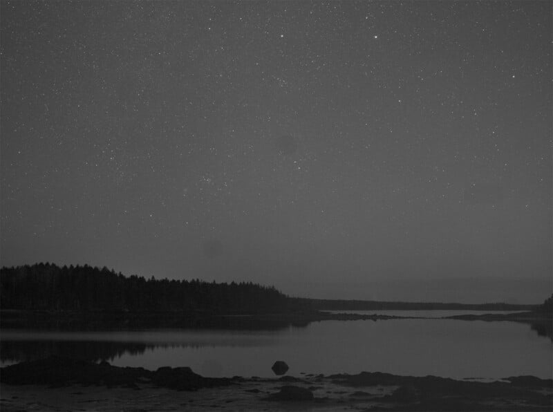 A serene black and white image depicting a calm lake reflecting the starry night sky. In the distance, a line of trees outlines the horizon, creating a tranquil and timeless atmosphere.