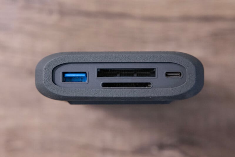 Close-up view of an external hard drive showing various ports on the side. The ports include a blue USB 3.0 port, two SD card slots, and a USB-C port. The background is a blurred, wooden surface.