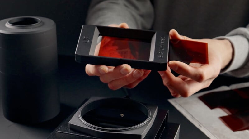 A person is holding photographic filters, preparing to place a red filter into a square holder. A black cylindrical container is nearby, and additional filters are on the table. The background is dark, emphasizing the process of photography setup.