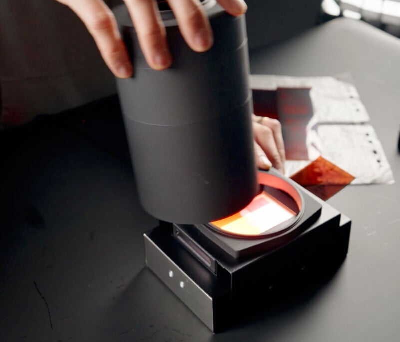 A person uses a black cylindrical film scanner on a dark table to digitize photographic negatives. The scanner glows with a red light, and various film strips are placed nearby.