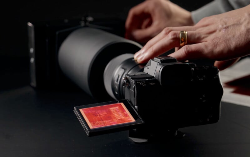 A person adjusts a black camera with a large lens. The camera's display screen is flipped out, showing a red-tinted image. The person's hand, adorned with a gold ring, is adjusting dials on the camera. The background is dimly lit.