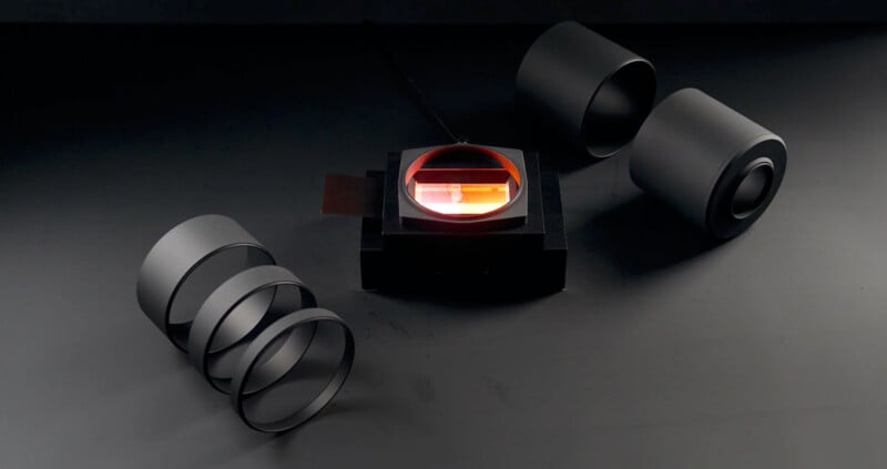 A set of black cylindrical components surrounds a central illuminated lens with a reddish glow on a dark surface. The arrangement suggests a focus on optics or technology.