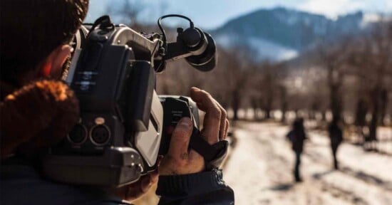 A person films with a handheld video camera on a snowy path surrounded by trees. Two figures walk in the distance toward a mountain under a clear blue sky.