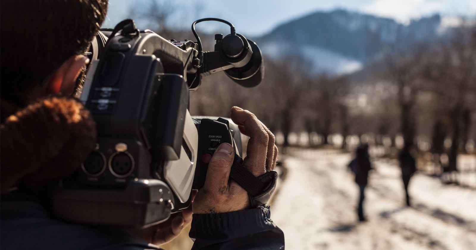 A person films with a handheld video camera on a snowy path surrounded by trees. Two figures walk in the distance toward a mountain under a clear blue sky.