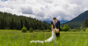 A couple in wedding attire embraces in a lush green meadow surrounded by tall trees and mountains under a cloudy sky. The bride holds a bouquet of yellow flowers, and they share a tender moment.