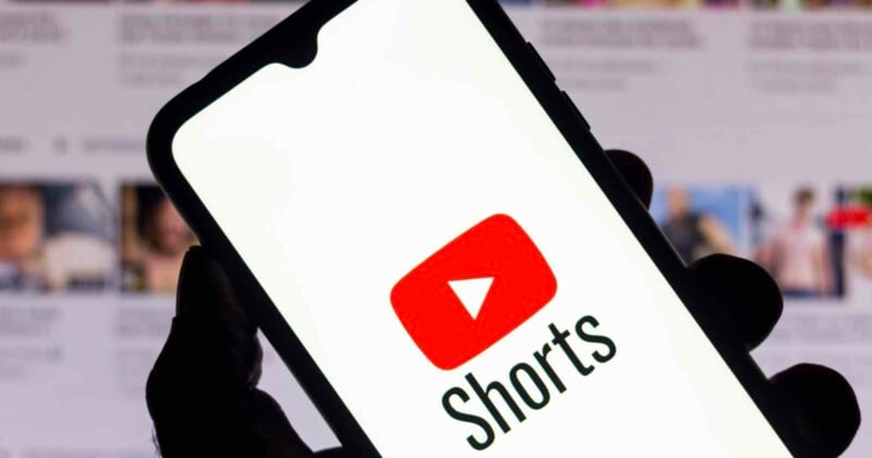 A hand holds a smartphone displaying the YouTube logo with the word "Shorts" below it. The background shows a blurred screen with thumbnails of multiple videos.