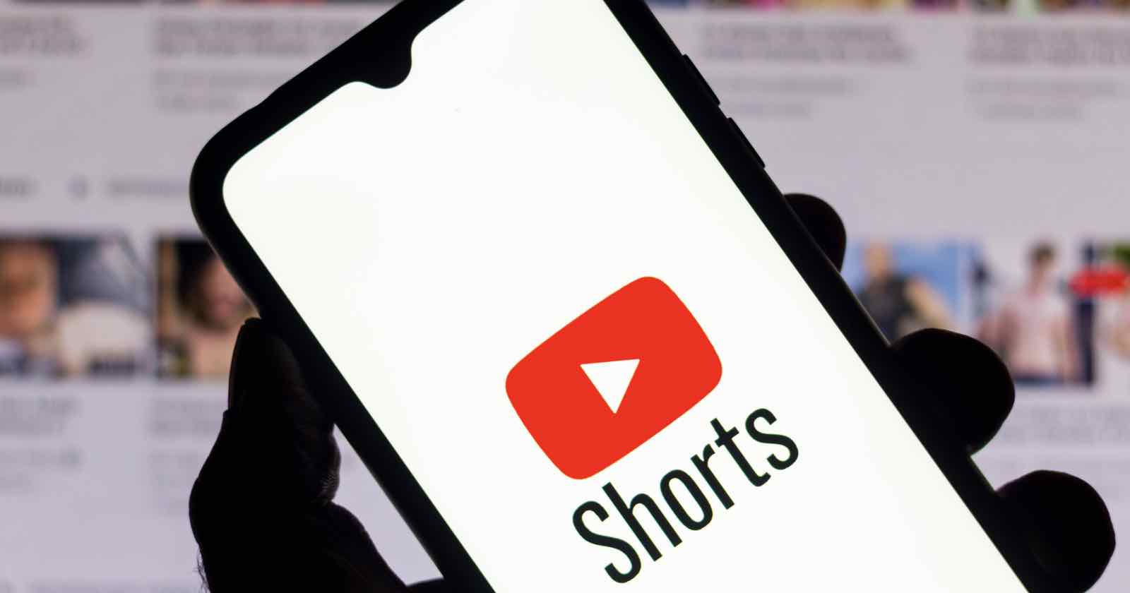 A hand holds a smartphone displaying the YouTube logo with the word "Shorts" below it. The background shows a blurred screen with thumbnails of multiple videos.