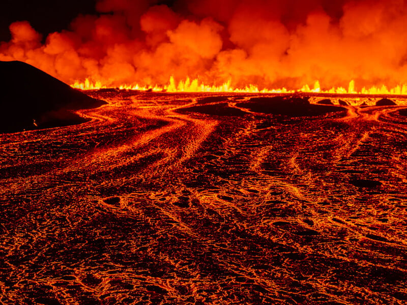 A vivid image of a lava field with bright orange and red molten rock flowing on the ground. Fiery flames and smoke rise into the night sky, creating a dramatic and intense scene of volcanic activity.