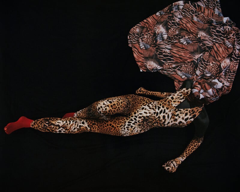 A person is lying on a dark surface, fully covered in body paint and fabric with leopard prints. The fabric slightly obscures their face while their red-painted foot is visible. The artistic arrangement blends with the background.