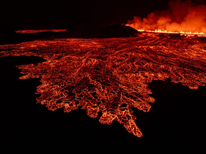 Aerial view of an expansive lava field glowing with bright orange and red molten rock. Smoke rises in the background, while fiery rivers of lava spread out over the dark landscape, creating a striking contrast against the night sky.