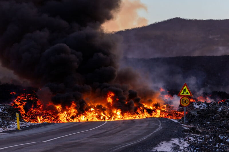 A road is blocked by a flow of lava, spewing flames and thick black smoke. A traffic sign indicates a speed limit of 50 and warns of construction. Mountains are visible in the background under a partly cloudy sky.