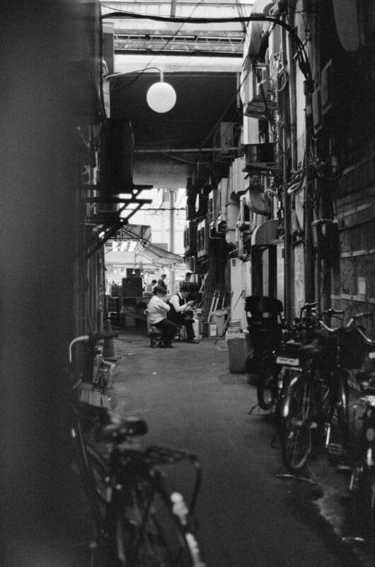 Photo en noir et blanc d’une ruelle urbaine étroite avec des vélos garés sur les côtés. Deux personnes sont assises ensemble au loin, sous un éclairage tamisé. La scène véhicule une atmosphère urbaine intime et calme.