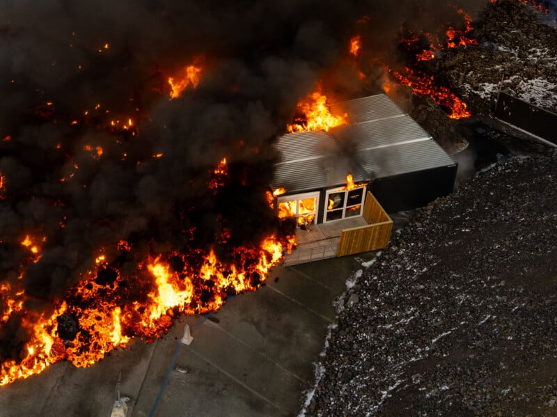 Aerial view of a large fire engulfing a building and surrounding areas with thick black smoke. The flames are intense and widespread, consuming a nearby structure, with charred debris on the ground.