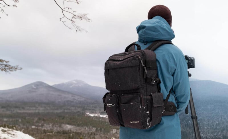 A person in a blue jacket stands on a snowy hill, facing away, looking at a mountain landscape. They have a large black backpack and are using a camera on a tripod. Bare tree branches frame the overcast sky.