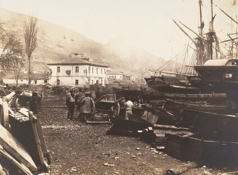 A sepia-toned photograph depicting a cluttered shipyard with several people standing among debris and ship parts. There are two large ships docked on the right and a white building in the background against a hilly terrain.