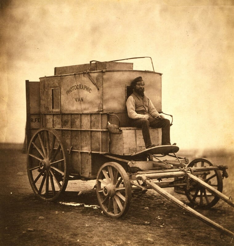 A sepia-toned image of a man sitting on a vintage photographic van, which is a covered wagon with large wooden wheels, labeled "Photographic Van." The van is parked on a barren landscape, evoking a historical setting.