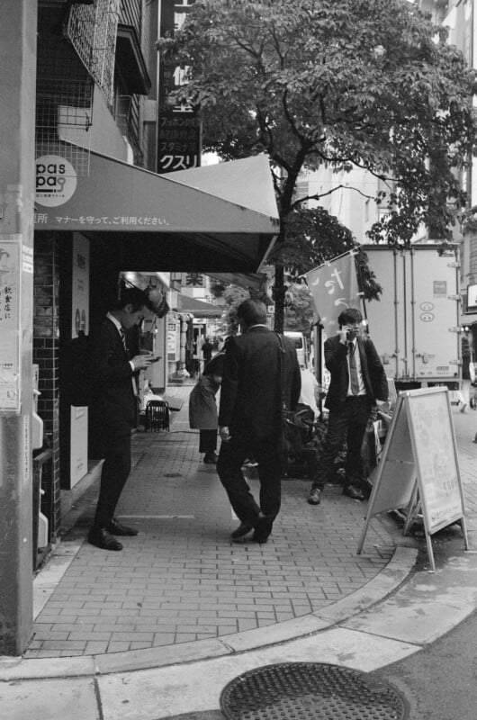 Une scène de rue en noir et blanc montre plusieurs personnes en tenue professionnelle debout près d’une devanture de magasin et d’un arbre. Ils sont en conversation ou regardent autour d’eux. Il y a des panneaux et un panneau sandwich sur le trottoir.